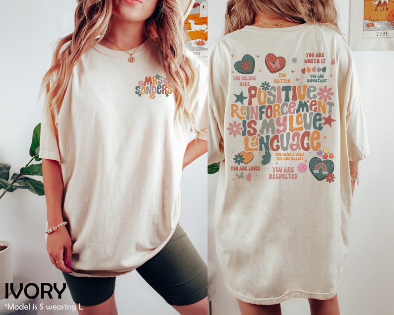 Pode incluir: Camiseta oversized de cor marfim com um design floral nas costas que diz "Refor&ccedil;o positivo &eacute; a minha linguagem de amor". A frente da camiseta tem um pequeno design floral com o texto "Mrs. Sanders".