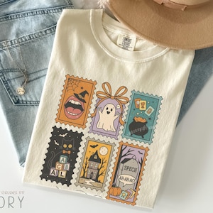 May include: Ivory-colored Comfort Colors t-shirt featuring a collection of Halloween-themed postage stamp designs. The stamps showcase a witch's mouth, a ghost, a black cat, a haunted house, and a tombstone. The shirt is paired with blue jeans and a tan hat.