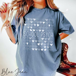 Pode incluir: Camiseta azul jeans com o texto branco "SCHOOL PSYCH", rodeado por um quadrado de cora&ccedil;&otilde;es brancos. A camisa &eacute; de cor confort&aacute;vel e n&atilde;o &eacute; uma camisa grande.