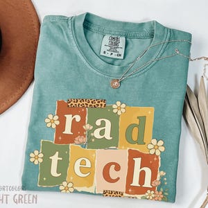 May include: A light green Comfort Colors t-shirt with the words "rad tech" in a retro design. The design features floral accents and a leopard print tape detail. A silver necklace with a pendant is draped across the shirt.
