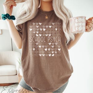 Pode incluir: Camiseta marrom com a frase "SCHOOL PSYCH" em branco, rodeada por uma grade de ilustra&ccedil;&otilde;es de cora&ccedil;&otilde;es brancos. A camiseta &eacute; de cor neutra e tem um corte casual e descontra&iacute;do. Uma caneca com a frase "COOL MOMS CLUB" &eacute; segurada na m&atilde;o.