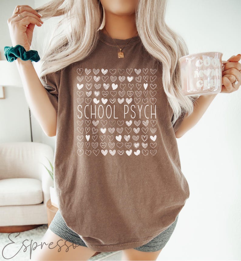Pode incluir: Camiseta marrom com a frase "SCHOOL PSYCH" em branco, rodeada por uma grade de ilustra&ccedil;&otilde;es de cora&ccedil;&otilde;es brancos. A camiseta &eacute; de cor neutra e tem um corte casual e descontra&iacute;do. Uma caneca com a frase "COOL MOMS CLUB" &eacute; segurada na m&atilde;o.