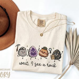 May include: An ivory t-shirt featuring a graphic of four cartoon rocks with faces, including one with a headlamp and pickaxe, an amethyst geode, one wearing a cap, and one holding a magnifying glass and a notebook labeled Field Notes, with the text wait, I see a rock.