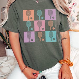 May include: An olive green t-shirt featuring a grid design of pickleball paddles and balls in various colours. The squares are orange, teal, pink, and purple. The text "COMFORT COLORS" and "Moss" are printed at the bottom of the shirt.