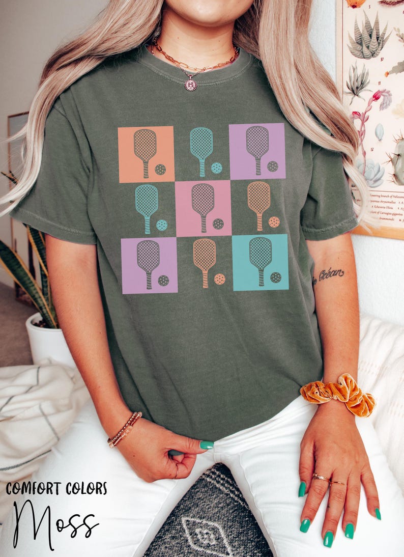 May include: An olive green t-shirt featuring a grid design of pickleball paddles and balls in various colours. The squares are orange, teal, pink, and purple. The text "COMFORT COLORS" and "Moss" are printed at the bottom of the shirt.