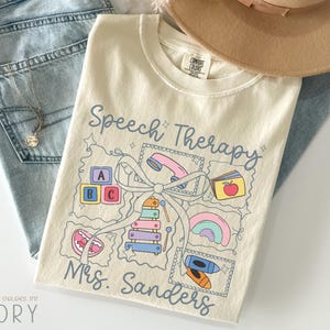 May include: Ivory-colored t-shirt with the words "Speech Therapy" and "Mrs. Sanders" in blue script. The shirt features a graphic of speech therapy tools, including a telephone, ABC blocks, and a xylophone. A tan hat and blue jeans are in the background.