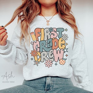 May include: A gray sweatshirt with the text "First Grade Crew" in colorful, retro-style lettering. The design includes a pencil, stars, and flowers.