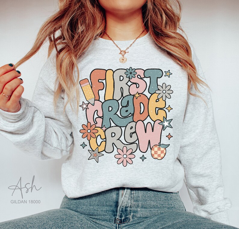 May include: A gray sweatshirt with the text "First Grade Crew" in colorful, retro-style lettering. The design includes a pencil, stars, and flowers.