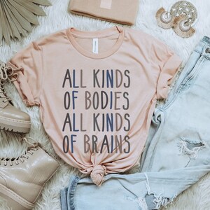 Może przedstawiać: Jasnor&oacute;żowy t-shirt z napisem "ALL KINDS OF BODIES ALL KINDS OF BRAINS" w niebieskich i szarych literach. Koszulka leży na białym, puszystym podłożu z parą jasnobrązowych but&oacute;w, parą jasnoniebieskich dżins&oacute;w i jasnobrązową kopert&oacute;wką.
