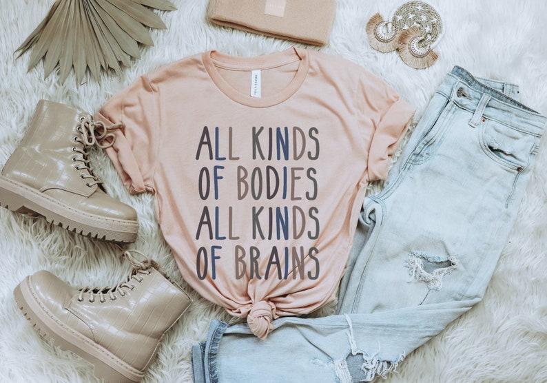 Może przedstawiać: Jasnor&oacute;żowy t-shirt z napisem "ALL KINDS OF BODIES ALL KINDS OF BRAINS" w niebieskich i szarych literach. Koszulka leży na białym, puszystym podłożu z parą jasnobrązowych but&oacute;w, parą jasnoniebieskich dżins&oacute;w i jasnobrązową kopert&oacute;wką.