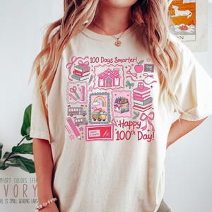 May include: Ivory-colored t-shirt with a colorful graphic celebrating the 100th day of school. The design includes school buses, books, pencils, and the text "100 Days Smarter!" and "Happy 100th Day!" in pink.