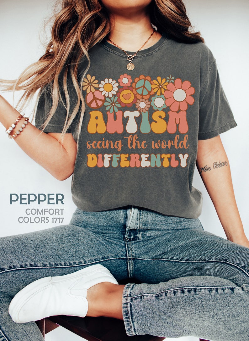 May include: A gray t-shirt with a colorful graphic design featuring flowers, peace signs, and the text "AUTISM seeing the world DIFFERENTLY".