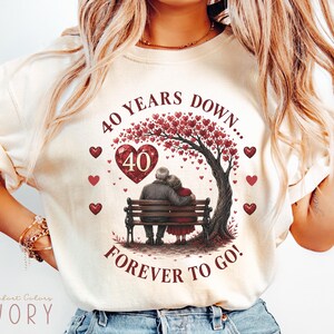 Könnte beinhalten: Ein elfenbeinfarbenes T-Shirt mit einer Grafik eines älteren Paares, das auf einer Bank unter einem Baum mit herzförmigen roten Blättern sitzt. Der Text lautet 40 YEARS DOWN... FOREVER TO GO! mit einem großen roten Herz und der Zahl 40.