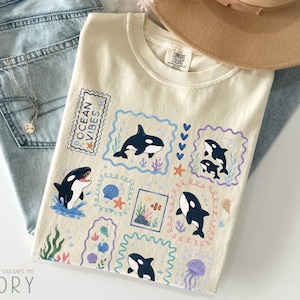 May include: An ivory t-shirt featuring a graphic print of multiple orca whales, seashells, coral, and a jellyfish arranged in rectangular frames. The text "OCEAN VIBES" is printed in a vertical stamp-like box. The design uses blue, purple, and orange.