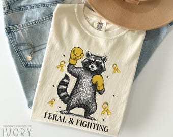 May include: Ivory-colored t-shirt featuring a raccoon wearing sunglasses and boxing gloves. The raccoon is surrounded by yellow ribbons and stars, with the words "FERAL & FIGHTING" printed below. The shirt is on top of blue jeans.