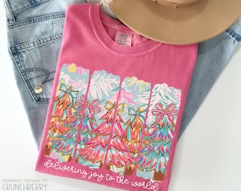 May include: A pink Comfort Colors t-shirt with a colorful Christmas tree design. The graphic features trees with bows and the text "delivering joy to the world." A tan hat and blue jeans are also in the image.