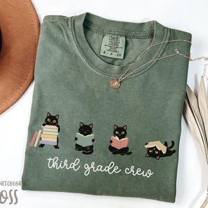 Puede incluir: Una camiseta de manga corta color verde musgo con un gráfico de cuatro gatos negros leyendo libros y el texto blanco "third grade crew". La camiseta tiene una etiqueta de la marca Comfort Colors en el cuello, indicando talla S-CH y 100% algodón.