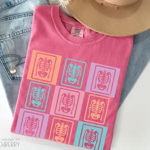 Puede incluir: Una camiseta rosa con un patrón de cuadrícula de diseños de esqueletos coloridos. La camiseta está doblada sobre unos vaqueros azules, con un sombrero marrón encima. El texto "COMFORT COLORS 1717 CRUNCHBERRY" es visible.