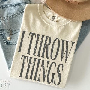 May include: Ivory-colored t-shirt with the text "I THROW THINGS" in dark gray, distressed lettering. The shirt is laid on top of light blue jeans and a tan hat. A silver necklace with a pendant is visible.