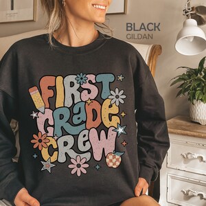 May include: Black sweatshirt with a retro-style graphic that says "First Grade Crew" in colorful letters. The graphic also includes a pencil, flowers, stars, and an apple.