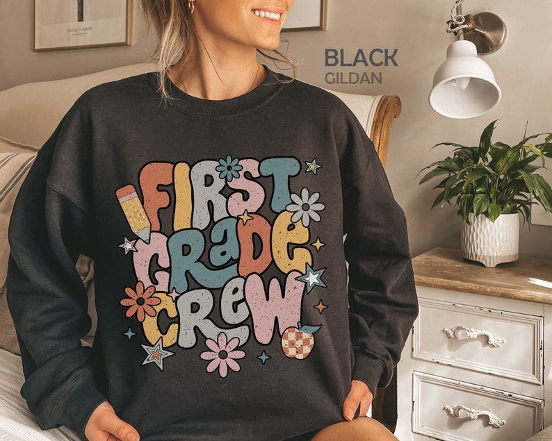 May include: Black sweatshirt with a retro-style graphic that says "First Grade Crew" in colorful letters. The graphic also includes a pencil, flowers, stars, and an apple.