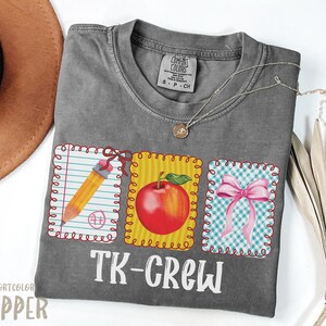 May include: A gray Comfort Colors t-shirt with a colorful graphic. The graphic includes a pencil, a red apple, and a pink bow, each in a decorative frame. The text "TK-CREW" is printed below the graphics. A gold necklace is draped across the shirt.