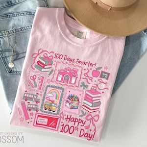 May include: A light pink t-shirt with a colorful graphic celebrating the 100th day of school. The design includes books, a school bus, crayons, and the text "100 Days Smarter!" and "Happy 100th Day!" The shirt is paired with blue jeans and a tan hat.