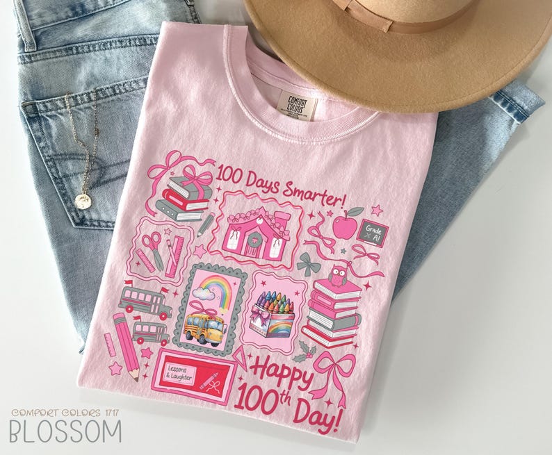 May include: A light pink t-shirt with a colorful graphic celebrating the 100th day of school. The design includes books, a school bus, crayons, and the text "100 Days Smarter!" and "Happy 100th Day!" The shirt is paired with blue jeans and a tan hat.