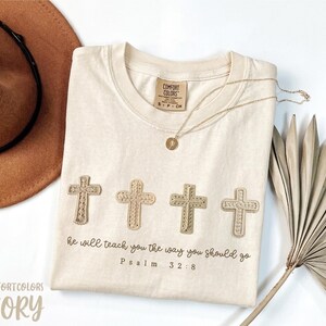 Peut inclure: T-shirt ivoire avec quatre croix brodées et le texte « il vous enseignera le chemin que vous devez suivre Psaume 32:8 ». Un collier doré et un chapeau marron sont également visibles.