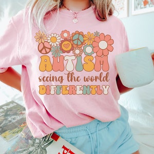 May include: Pink t-shirt with a retro-style graphic design featuring flowers, peace signs, and the text "AUTISM seeing the world DIFFERENTLY".