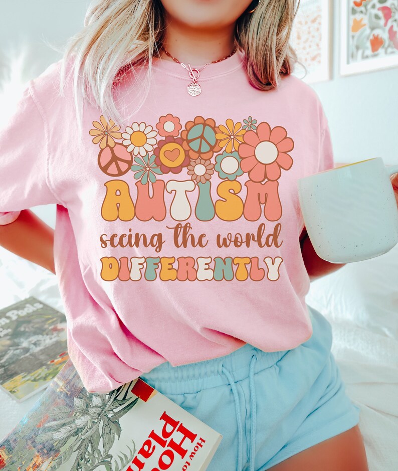 May include: Pink t-shirt with a retro-style graphic design featuring flowers, peace signs, and the text "AUTISM seeing the world DIFFERENTLY".