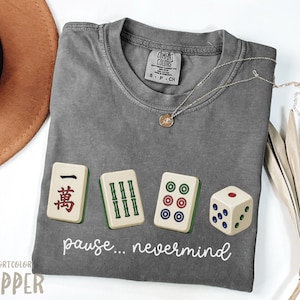 Puede incluir: Una camiseta de manga corta gris carbón con un gráfico de tres fichas de mahjong y un dado de seis caras. Debajo de los gráficos, el texto "pause... nevermind" aparece en letra cursiva blanca. La camiseta tiene cuello redondo y una etiqueta.