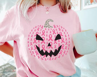 Op de afbeelding: Roze shirt met lange mouwen met een pompoenontwerp gemaakt van roze linten. De pompoen heeft zwarte ogen en een grillige mond. Het shirt is lichtroze van kleur en heeft een casual stijl.