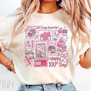 May include: Ivory-colored t-shirt with a colorful graphic celebrating the 100th day of school. The design includes school-themed illustrations such as books, pencils, a school bus, and the text "100 Days Smarter!" and "Happy 100th Day!"
