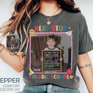 Puede incluir: Camiseta gris oscuro con una foto de una niña pequeña sosteniendo una pizarra que dice "My First Day of Preschool". La camiseta tiene las palabras "Next Stop Preschool Middle School" y el nombre "Sarah".