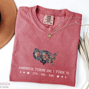 May include: A crimson Comfort Colors t-shirt with a graphic of the United States map made of flags. The text "AMERICA TURNS 250, I TURN 70" is printed below the map, with the years 1776-1956-2026. A gold necklace with a pendant is on the shirt.