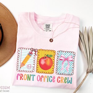 May include: A light pink t-shirt with the words "FRONT OFFICE CREW" in colorful letters. The shirt features three framed graphics: a pencil, a red apple, and a pink bow. A gold necklace is draped on the shirt.