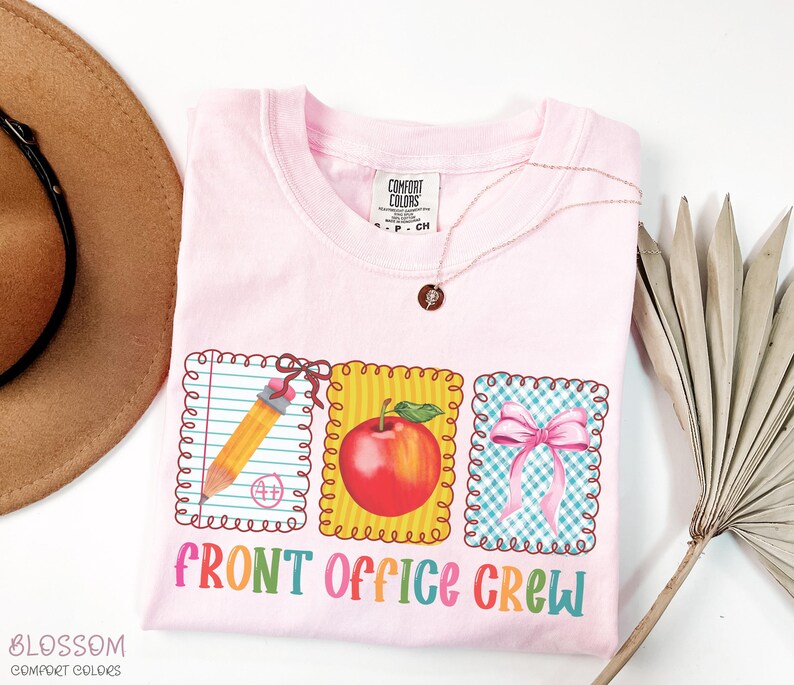 May include: A light pink t-shirt with the words "FRONT OFFICE CREW" in colorful letters. The shirt features three framed graphics: a pencil, a red apple, and a pink bow. A gold necklace is draped on the shirt.