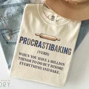 May include: Ivory-colored t-shirt with the text "PROCRASTIBAKING" and a rolling pin graphic. The shirt's text explains the meaning of the word. The shirt is paired with blue jeans and a tan hat.