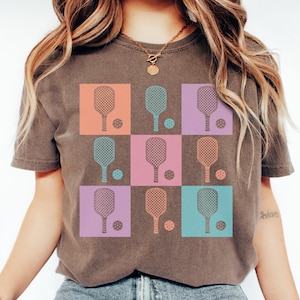 May include: A brown t-shirt with a grid design featuring pickleball paddles and balls. The squares are in orange, blue, purple, and pink. The pickleball paddles and balls are in matching colours.