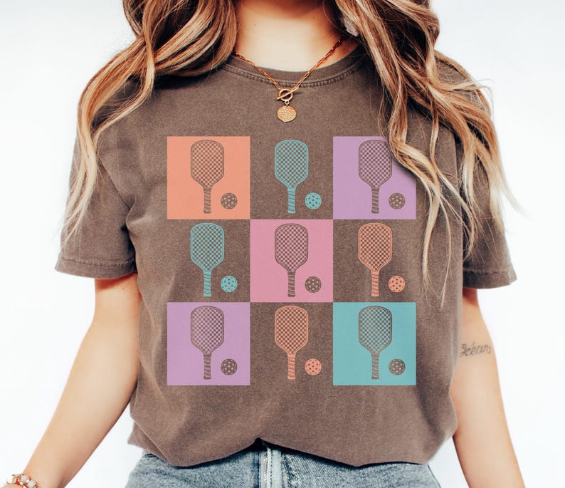 May include: A brown t-shirt with a grid design featuring pickleball paddles and balls. The squares are in orange, blue, purple, and pink. The pickleball paddles and balls are in matching colours.