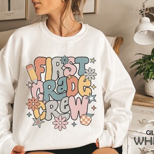 May include: White sweatshirt with a colorful graphic that says "First Grade Crew" with a pencil, stars, and flowers. The text is in a retro font style.