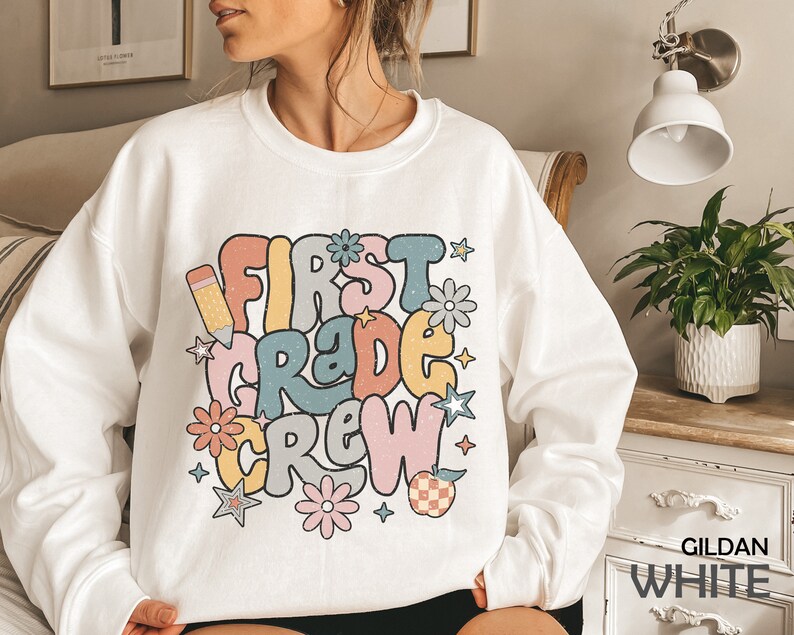 May include: White sweatshirt with a colorful graphic that says "First Grade Crew" with a pencil, stars, and flowers. The text is in a retro font style.