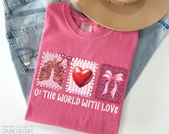 Puede incluir: Una camiseta rosa Comfort Colors con el texto "O2 The World With Love". La camiseta presenta tres diseños cuadrados: pulmones florales, un corazón rojo y una caja de regalo con un lazo rosa. Un sombrero beige y unos vaqueros azules están en el fondo.