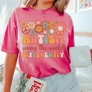 May include: A pink t-shirt with a retro-style graphic design featuring flowers, peace signs, and the text "AUTISM seeing the world DIFFERENTLY".