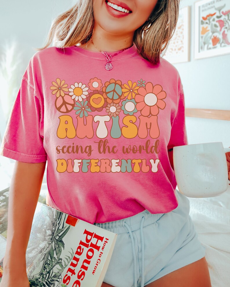May include: A pink t-shirt with a retro-style graphic design featuring flowers, peace signs, and the text "AUTISM seeing the world DIFFERENTLY".