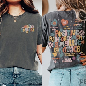 Pode incluir: Uma camiseta cinza escura com um gr&aacute;fico colorido nas costas que diz "Positive Reinforcement is my love language". A frente da camiseta tem um gr&aacute;fico menor que diz "Mrs. Sanders".
