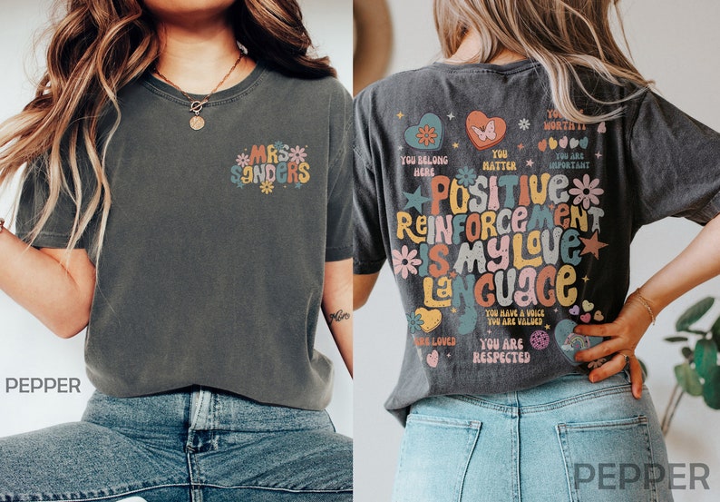 Pode incluir: Uma camiseta cinza escura com um gr&aacute;fico colorido nas costas que diz "Positive Reinforcement is my love language". A frente da camiseta tem um gr&aacute;fico menor que diz "Mrs. Sanders".