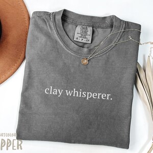 Puede incluir: Camiseta gris con el texto "clay whisperer." impreso en blanco en la parte delantera. La camiseta está hecha de una tela suave y cómoda.