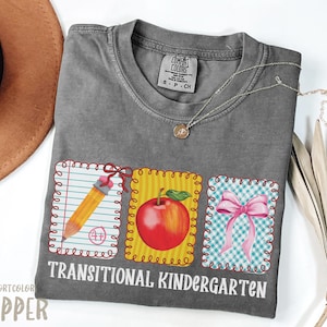 May include: Gray Comfort Colors t-shirt with a pencil, apple, and bow design. The shirt has the words "Transitional Kindergarten" printed on it. A gold necklace is draped across the shirt.